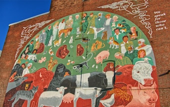 A mural painted on a brick wall depicting numerous people, animals, and nature elements. The image contains humans dressed in various clothes, standing and interacting with a diverse range of animals, including dogs, cats, cows, sheep, birds, and wild animals. The background features green and earthy tones, and there is text on the right side that reads, 'We speak for those who can't.'