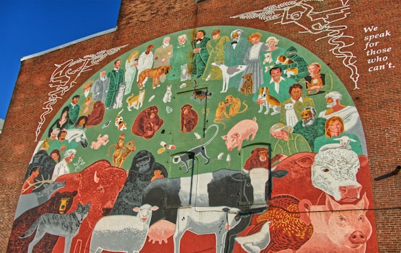 A mural painted on a brick wall depicting numerous people, animals, and nature elements. The image contains humans dressed in various clothes, standing and interacting with a diverse range of animals, including dogs, cats, cows, sheep, birds, and wild animals. The background features green and earthy tones, and there is text on the right side that reads, 'We speak for those who can't.'