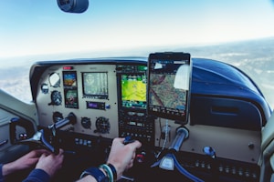 The interior of an airplane cockpit with various controls, screens, and gauges displayed. A person is holding onto the control yoke, while a tablet mounted on the dashboard shows a digital map.