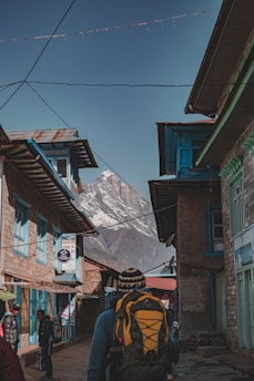 A person with a backpack and a knitted hat walks through a narrow street lined with traditional stone and wooden buildings. The buildings have a rustic appearance and are painted in shades of blue. In the background, a towering snow-capped mountain rises against a clear blue sky. Additional people are seen walking along the street.