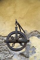 An old, rusted iron pulley is mounted on a weathered wall. The pulley features a circular wheel with a sturdy metal frame and visible chains. The wall behind it has peeling paint and rough textures, with shades of yellow and gray.