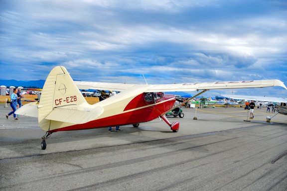A vintage red and cream aircraft is parked on an airstrip. The scene is bustling with people walking around, possibly at an airshow or exhibition. Several other small aircraft are lined up in the background under a cloudy sky.