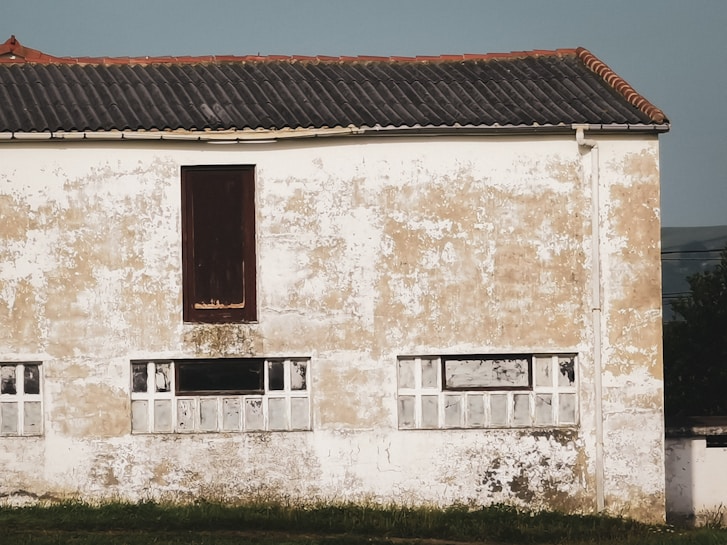 A weathered plaster exterior facade with patches of peeling paint is seen on a building, featuring a dark brown boarded-up window and several smaller, partially broken or filled windows beneath. The roof is made of dark clay tiles and there is a strip of grass in the foreground.