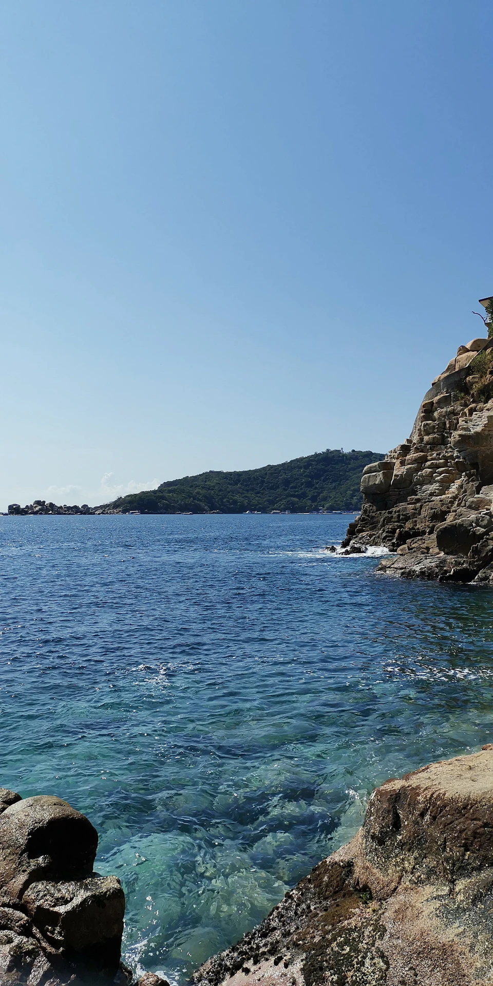 The image depicts a serene coastal scene with a rocky cliff on one side and a deep blue ocean on the other, creating a striking contrast between the rugged terrain and the tranquil water body. The sky above is clear and blue, suggesting a bright and sunny day. In the distance, there are several small buildings visible near the shoreline, adding to the picturesque landscape.
