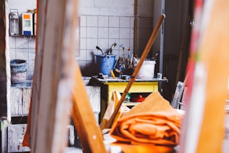 A cluttered artist's studio featuring a variety of painting tools and materials. Paintbrushes are stored in containers on a table alongside a yellow shelf with additional art supplies. There are paint cans and various items scattered around. The setting is colorful and has a rustic, creative atmosphere.