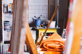 A cluttered artist's studio featuring a variety of painting tools and materials. Paintbrushes are stored in containers on a table alongside a yellow shelf with additional art supplies. There are paint cans and various items scattered around. The setting is colorful and has a rustic, creative atmosphere.