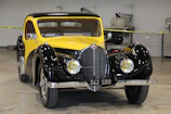 A vintage car with a glossy black and yellow exterior, showcasing a classic design with rounded fenders and chrome accents. The car is parked indoors, surrounded by caution tape and various automotive tools in the background, indicating it might be part of a workshop or exhibition setting.