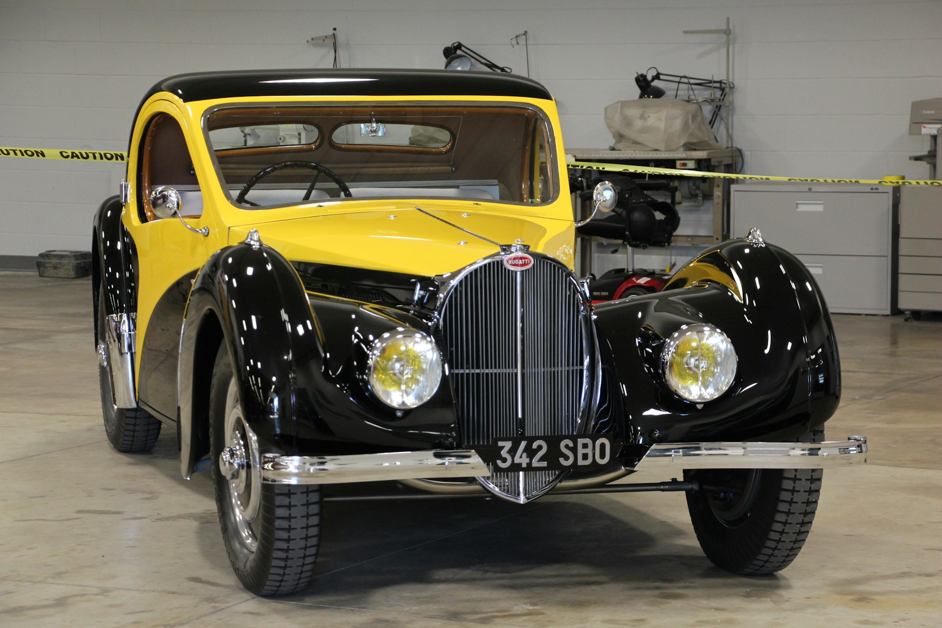 A vintage car with a glossy black and yellow exterior, showcasing a classic design with rounded fenders and chrome accents. The car is parked indoors, surrounded by caution tape and various automotive tools in the background, indicating it might be part of a workshop or exhibition setting.