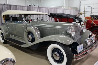 A classic vintage car with a sleek, shiny gray body, white-wall tires, and chrome detailing is parked indoors. It has a convertible top and the license plate indicates it is from Ontario. The car is part of a collection, with other vintage vehicles visible in the background.