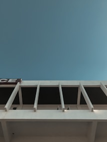 The image features a modern architectural structure with clean, linear design elements against a clear blue sky. Vertical beams extend from the building, creating a rhythmic pattern.