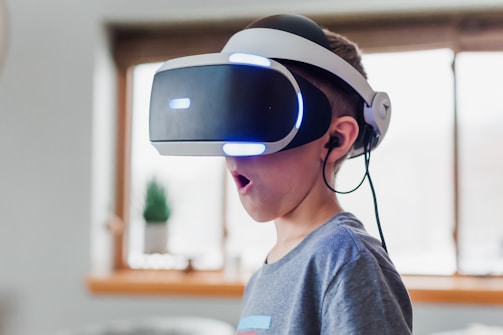 A young boy wearing a virtual reality headset appears to be engaged and surprised. The environment is indoors, with soft lighting and a blurred background. The boy is wearing a casual grey T-shirt, and the headset has glowing blue lights.