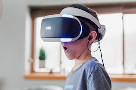 A young boy wearing a virtual reality headset appears to be engaged and surprised. The environment is indoors, with soft lighting and a blurred background. The boy is wearing a casual grey T-shirt, and the headset has glowing blue lights.