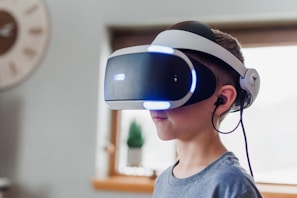 A young person wearing a virtual reality headset is standing indoors. The headset has blue lights and covers their eyes. Earbuds are attached, and the background includes a blurred window with some greenery visible.