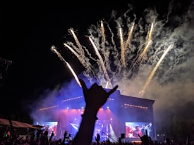 A nighttime music concert with fireworks exploding in the sky above a stage with large screens. The crowd's hands are raised, creating an energetic atmosphere. A silhouette of a hand making a rock gesture is prominent in the foreground.