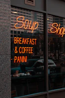 A storefront with a large window displaying a neon sign advertising soup, breakfast, coffee, and panini. Reflections of nearby buildings and trees can be seen in the glass. Inside, people are seated at tables enjoying food and drinks.