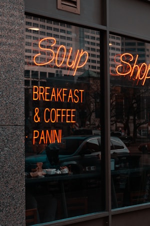 A storefront with a large window displaying a neon sign advertising soup, breakfast, coffee, and panini. Reflections of nearby buildings and trees can be seen in the glass. Inside, people are seated at tables enjoying food and drinks.