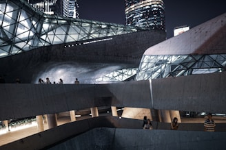 A modern architectural structure with geometric shapes and glass windows is illuminated at night. People are standing on different levels of the building. The environment is urban with a contrasting combination of concrete and glass, creating a futuristic ambiance.