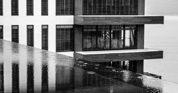 A black and white photograph showcasing a modern building with large glass windows and horizontal lines. The foreground features a polished surface with visible raindrops, indicating wetness. The juxtaposition of the reflective surface and the building's strong geometric lines creates a striking visual contrast.