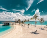 A stunning tropical beach scene with white sandy shores and turquoise waters. Palm trees sway gently in the breeze under a vibrant blue sky filled with fluffy clouds. A couple of beach huts can be seen along the sand, along with lounge chairs and a hammock. Small boats are visible on the water, adding to the idyllic setting.