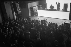 A packed crowd is gathered in a dimly lit venue, with a DJ performing on stage. The audience is engaged with arms raised, expressing excitement and enthusiasm. Two people stand behind a well-lit DJ booth, with one raising their arms, possibly encouraging the crowd. Large speakers and technical equipment are visible, suggesting this is a music concert or club event.