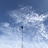A single streetlight pole extends upwards into a partly cloudy sky filled with white, wispy clouds against a blue background. The lamp post is slender and simple in design, contrasting with the natural patterns of the clouds.