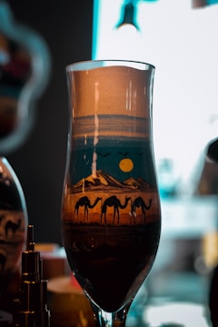 A glass container displaying an intricate sand art design featuring a desert scene with three camels walking in front of mountainous terrain under a yellow sun. The sand layers create a gradient of colors, enhancing the depiction of a desert landscape.