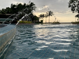 A tranquil swimming pool with clear water reflecting the sky. Palm trees line the background, silhouetted against a soft, cloudy sky. Water splashes into the pool from a fountain, capturing movement and sparkle. The setting appears serene, likely during a sunset or sunrise.