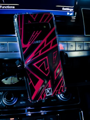 A smartphone with a vibrant, geometric patterned case is mounted on a car dashboard. The car's controls and part of the infotainment screen displaying 'Settings' and 'Functions' are visible, giving a tech-savvy and automotive ambiance.