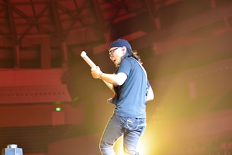 A musician energetically plays an electric guitar on stage. The environment is dimly lit, with dramatic stage lighting highlighting the performer. The backdrop shows an indoor venue with an industrial ceiling design.