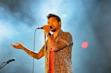 A male singer holds a microphone close to his mouth, wearing a floral shirt and displaying tattoos on his arm. The background is filled with smoke and colorful lights, creating a dynamic and engaging atmosphere.