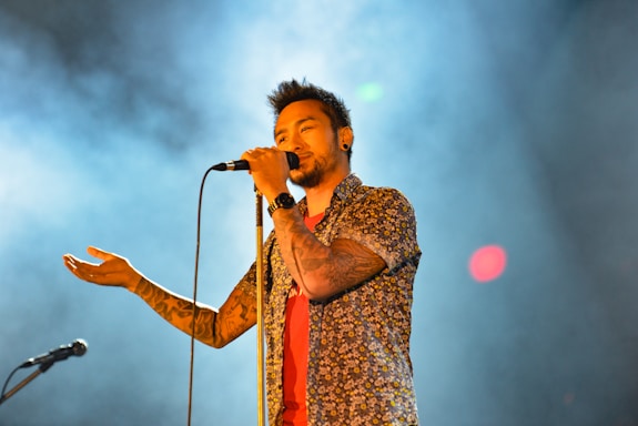 A male singer holds a microphone close to his mouth, wearing a floral shirt and displaying tattoos on his arm. The background is filled with smoke and colorful lights, creating a dynamic and engaging atmosphere.