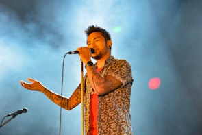 A male singer holds a microphone close to his mouth, wearing a floral shirt and displaying tattoos on his arm. The background is filled with smoke and colorful lights, creating a dynamic and engaging atmosphere.
