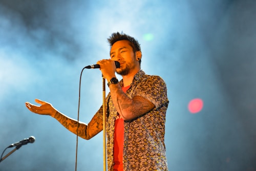 A male singer holds a microphone close to his mouth, wearing a floral shirt and displaying tattoos on his arm. The background is filled with smoke and colorful lights, creating a dynamic and engaging atmosphere.