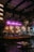 A cozy, dimly lit bar with a neon sign reading 'Bar Italiano' above a well-stocked shelf of bottles. Warm lighting hangs over the bar counter, and tables with chairs are arranged in the foreground. A person is working behind the counter, with glasses and bottles neatly organized.