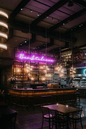 A cozy, dimly lit bar with a neon sign reading 'Bar Italiano' above a well-stocked shelf of bottles. Warm lighting hangs over the bar counter, and tables with chairs are arranged in the foreground. A person is working behind the counter, with glasses and bottles neatly organized.