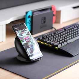 A smartphone is displayed on a wireless charging stand with its screen showing a social media feed. Nearby, a mechanical keyboard with RGB lighting sits on a dark mouse pad. In the background, a gaming console with blue and red controllers is visible on a desk.