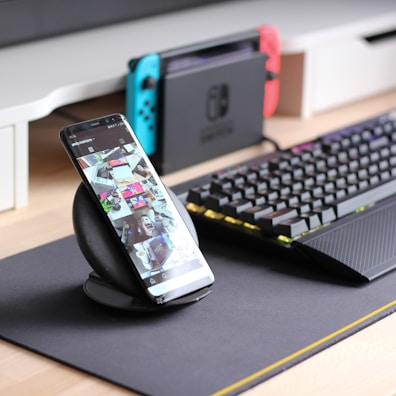 A smartphone is displayed on a wireless charging stand with its screen showing a social media feed. Nearby, a mechanical keyboard with RGB lighting sits on a dark mouse pad. In the background, a gaming console with blue and red controllers is visible on a desk.