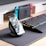 A smartphone is displayed on a wireless charging stand with its screen showing a social media feed. Nearby, a mechanical keyboard with RGB lighting sits on a dark mouse pad. In the background, a gaming console with blue and red controllers is visible on a desk.