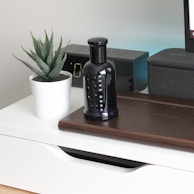 A sleek black perfume bottle is placed on a wooden surface beside a small potted plant with succulent leaves in a white pot. An electronic device with a digital display and a speaker are positioned in the background.