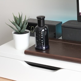 A sleek black perfume bottle is placed on a wooden surface beside a small potted plant with succulent leaves in a white pot. An electronic device with a digital display and a speaker are positioned in the background.