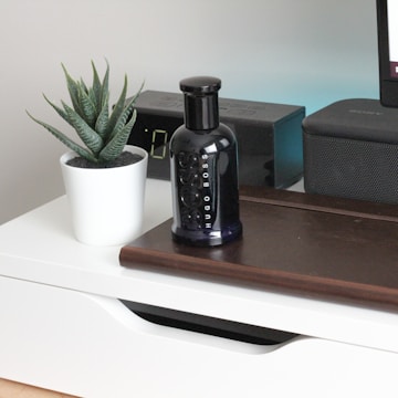 A sleek black perfume bottle is placed on a wooden surface beside a small potted plant with succulent leaves in a white pot. An electronic device with a digital display and a speaker are positioned in the background.