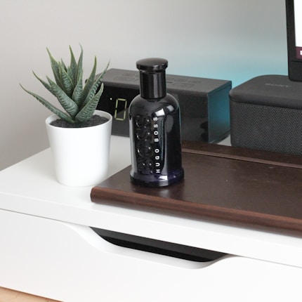A sleek black perfume bottle is placed on a wooden surface beside a small potted plant with succulent leaves in a white pot. An electronic device with a digital display and a speaker are positioned in the background.