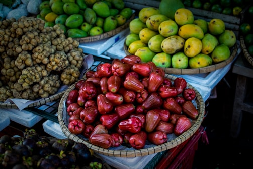 A variety of fruits are elegantly arranged in baskets at a market stall. At the forefront, there are glossy red rose apples piled in a woven basket. To the left, there is a bundle of small, round, beige-colored longans. In the background, ripe yellow and green mangoes fill another basket, creating a colorful contrast.