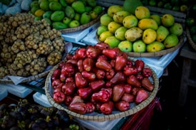 A variety of fruits are elegantly arranged in baskets at a market stall. At the forefront, there are glossy red rose apples piled in a woven basket. To the left, there is a bundle of small, round, beige-colored longans. In the background, ripe yellow and green mangoes fill another basket, creating a colorful contrast.