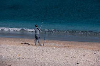 A person stands on a sandy beach, holding a fishing rod while facing the ocean. The waves gently crash onto the shore, and the clear blue water extends to the horizon. The beach is scattered with small rocks and shells.
