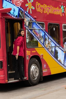 A person wearing a red jacket is stepping off a red city sightseeing tour bus. The bus features vibrant designs with photos of various city landmarks and the text 'City Sightseeing.' The background suggests an urban environment.