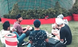 A group of young people with turbans sitting on a grassy area, engaging in conversation and enjoying fast food from 'Lord of the Fries'. They are surrounded by green plants in large red planters and there is a festive atmosphere indicated by the backdrop design.