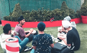 A group of young people with turbans sitting on a grassy area, engaging in conversation and enjoying fast food from 'Lord of the Fries'. They are surrounded by green plants in large red planters and there is a festive atmosphere indicated by the backdrop design.