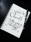 A spiral notebook with a motivational quote written in elegant, cursive handwriting. The phrase 'You're capable of amazing things' is prominently displayed. Accompanying the notebook is a black brush pen lying on the side. The notebook has a dotted background, adding to the artistic presentation.