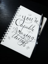 A spiral notebook with a motivational quote written in elegant, cursive handwriting. The phrase 'You're capable of amazing things' is prominently displayed. Accompanying the notebook is a black brush pen lying on the side. The notebook has a dotted background, adding to the artistic presentation.
